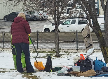 A woman speaks with an unsheltered person in a park setting with their belongings nearby