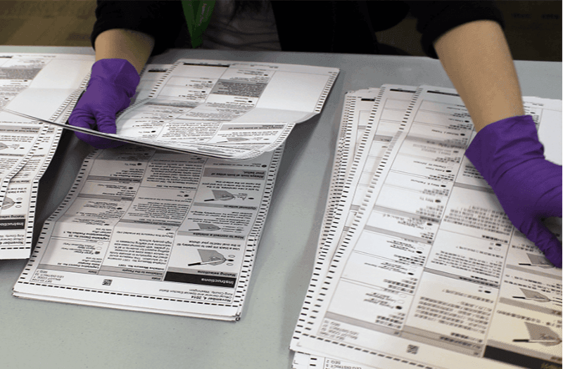 A person wearing purple gloves is handling multiple sheets of ballots.